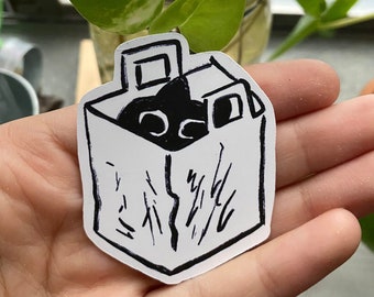 May include: A black and white sticker of a cat in a paper bag. The cat is peeking out of the bag with its eyes and nose visible. The bag has a handle and is drawn with a textured surface.