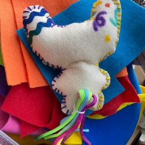 May include: A white felt butterfly with colorful embroidered details and a rainbow-colored string tail. The butterfly is sitting on a stack of colorful felt squares.