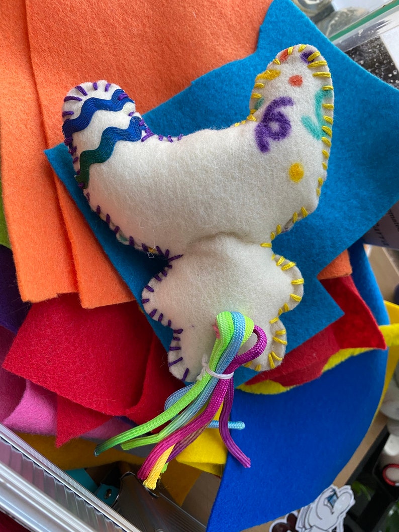 May include: A white felt butterfly with colorful embroidered details and a rainbow-colored string tail. The butterfly is sitting on a stack of colorful felt squares.
