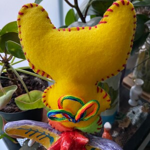 May include: A yellow felt rooster head with red stitching and a rainbow yarn bow. The rooster head is attached to a ceramic rooster figurine.