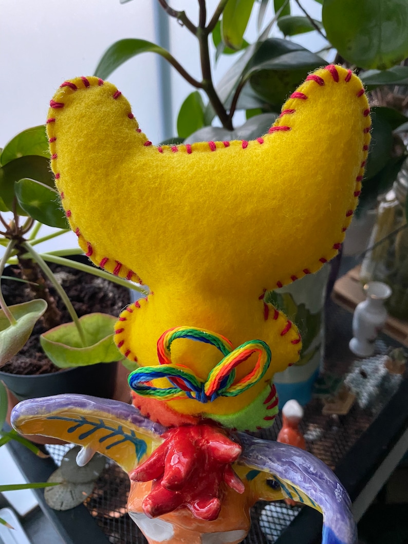 May include: A yellow felt rooster head with red stitching and a rainbow yarn bow. The rooster head is attached to a ceramic rooster figurine.