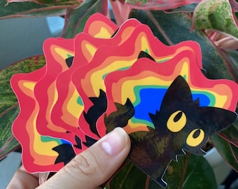 May include: A stack of rainbow-colored stickers featuring a black cat with yellow eyes. The cat is silhouetted against a blue and rainbow background.
