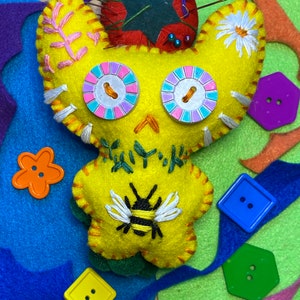 May include: A yellow felt toy with embroidered details, including a bee, flowers, and eyes. The toy is surrounded by colorful felt scraps and buttons.