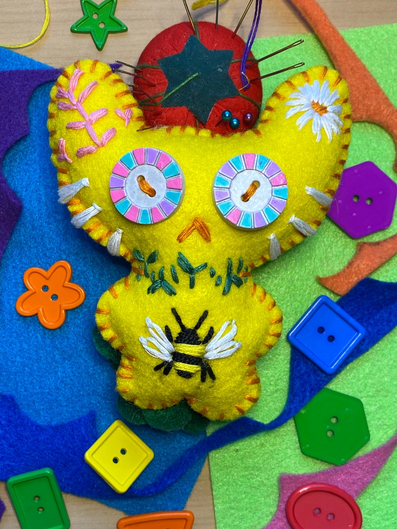 May include: A yellow felt toy with embroidered details, including a bee, flowers, and eyes. The toy is surrounded by colorful felt scraps and buttons.