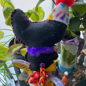 May include: A black felt toy with a purple fuzzy trim and a red and white party hat sits on a colorful ceramic bull figurine. The bull has a rainbow braided tail.