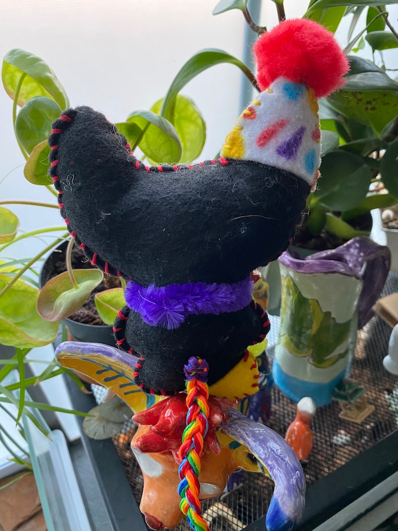 May include: A black felt toy with a purple fuzzy trim and a red and white party hat sits on a colorful ceramic bull figurine. The bull has a rainbow braided tail.