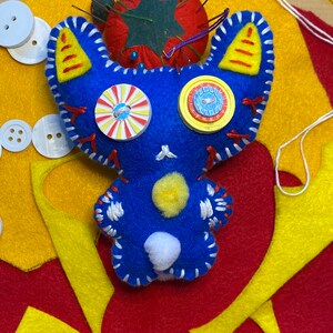 May include: Blue felt toy with white stitching, yellow ears, and white and yellow button eyes. The toy is surrounded by yellow and red felt scraps.