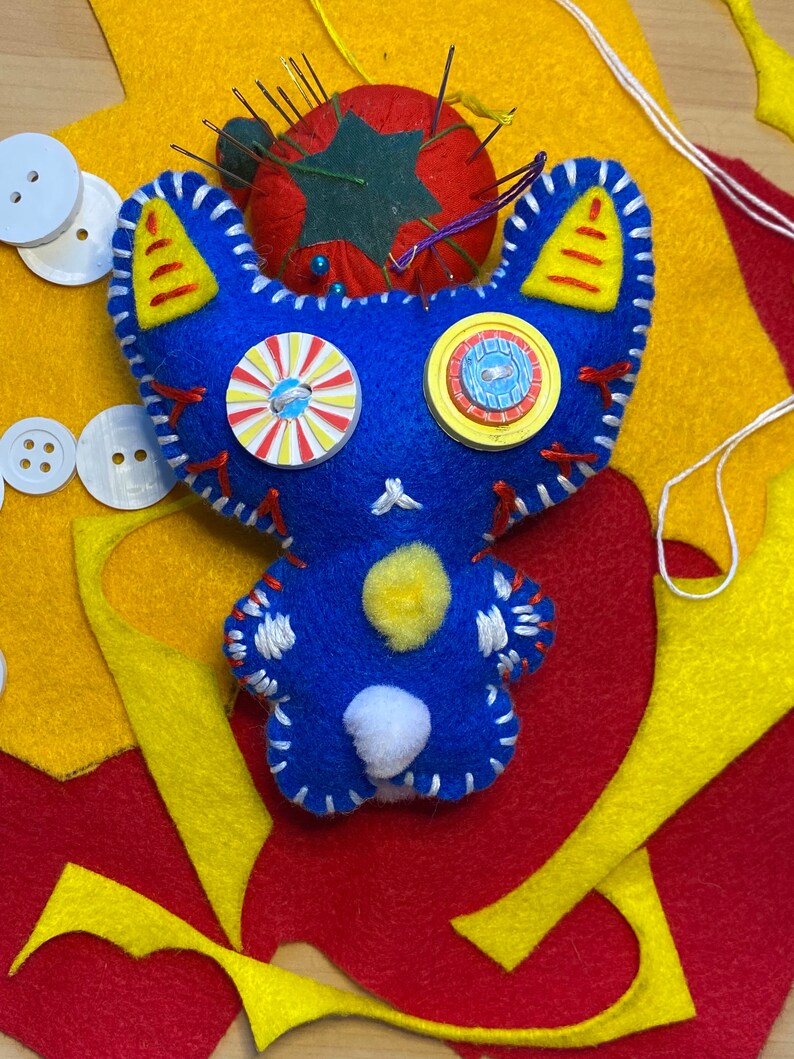 May include: Blue felt toy with white stitching, yellow ears, and white and yellow button eyes. The toy is surrounded by yellow and red felt scraps.