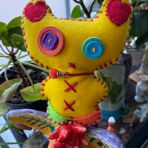 May include: A yellow felt toy with red heart patches, colorful buttons for eyes, and a red felt collar with a silver charm. The toy is sitting on a colorful ceramic figurine.