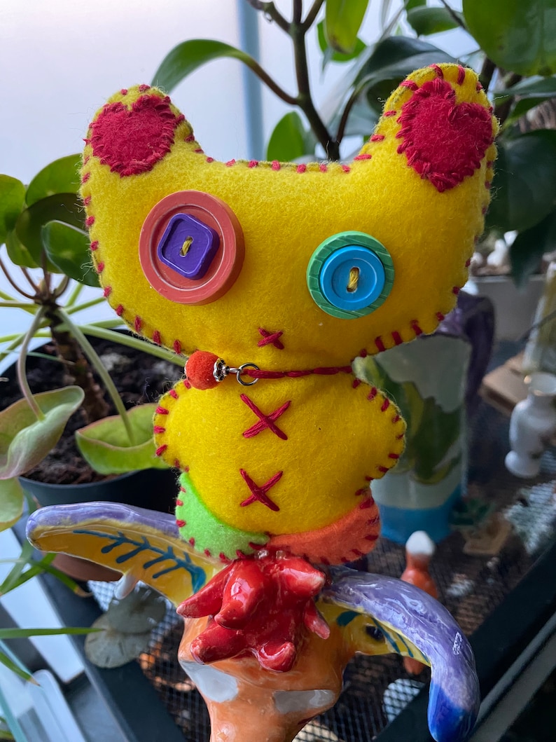 May include: A yellow felt toy with red heart patches, colorful buttons for eyes, and a red felt collar with a silver charm. The toy is sitting on a colorful ceramic figurine.