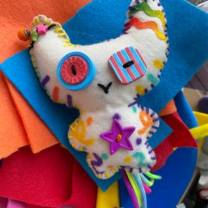 May include: A white felt toy with colorful embroidery and buttons. The toy has a star-shaped button, a striped button, and a round button with a red center. The toy has a long tail with colorful yarn strands.