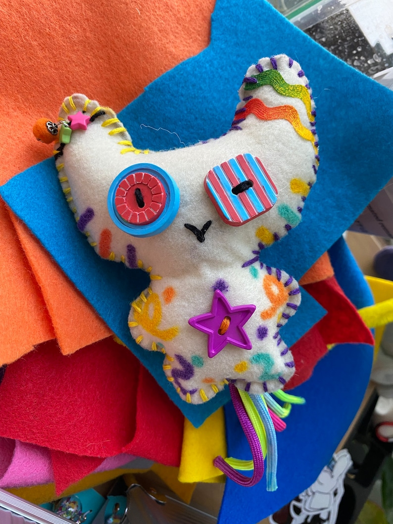 May include: A white felt toy with colorful embroidery and buttons. The toy has a star-shaped button, a striped button, and a round button with a red center. The toy has a long tail with colorful yarn strands.