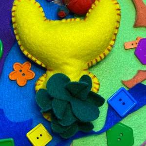 May include: A yellow felt craft project shaped like a flower with a green felt flower in the center. The project is surrounded by colorful felt scraps and buttons.