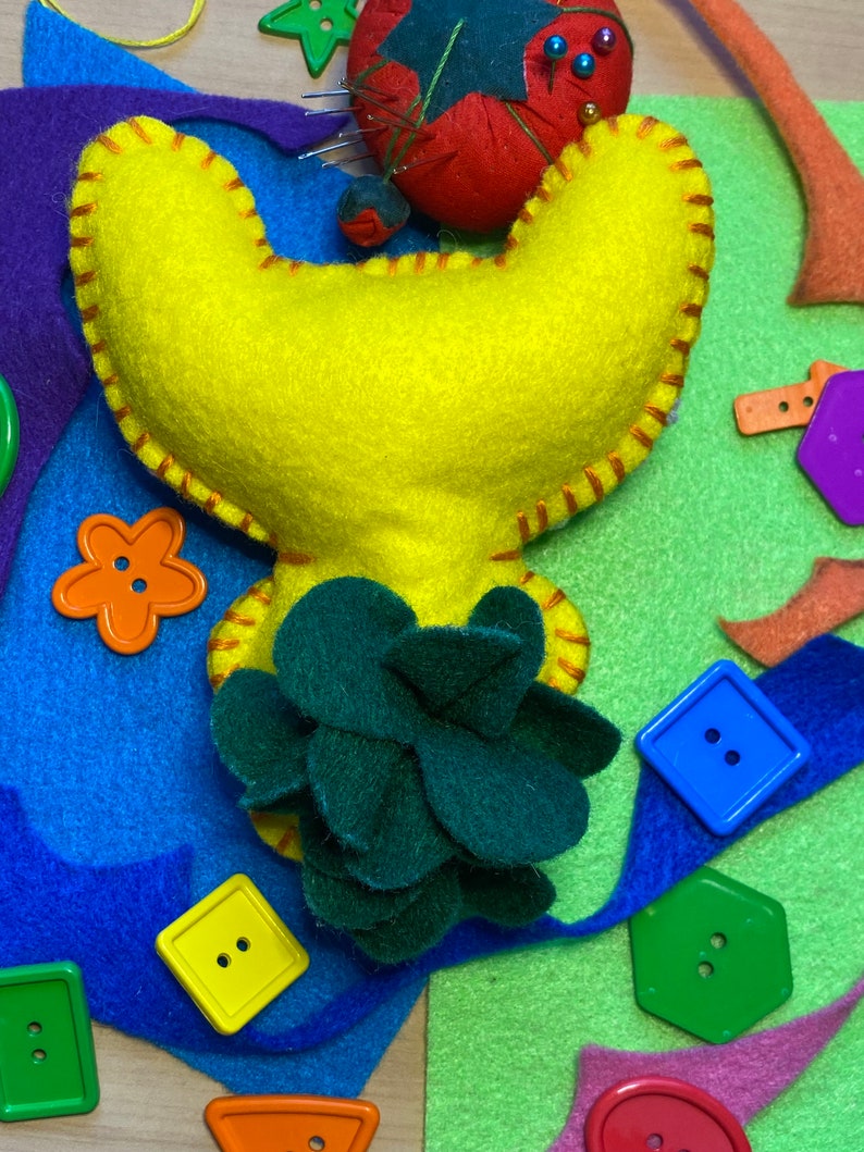 May include: A yellow felt craft project shaped like a flower with a green felt flower in the center. The project is surrounded by colorful felt scraps and buttons.