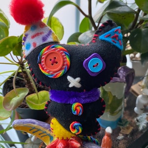 May include: A black felt cat toy with a rainbow candy cane design, purple fur trim, and colorful buttons. The cat is wearing a white and red party hat with a red pom-pom on top. The cat is perched on a colorful ceramic figurine.