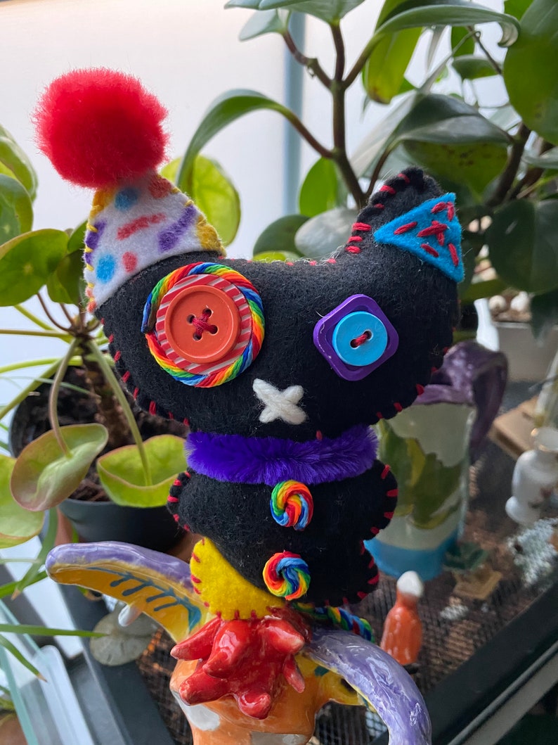 May include: A black felt cat toy with a rainbow candy cane design, purple fur trim, and colorful buttons. The cat is wearing a white and red party hat with a red pom-pom on top. The cat is perched on a colorful ceramic figurine.