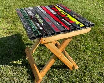 May include: A wooden folding stool with a seat made from hockey sticks. The sticks are arranged in rows and have various brand names, including "Bauer", "CCM", and "Warrior".