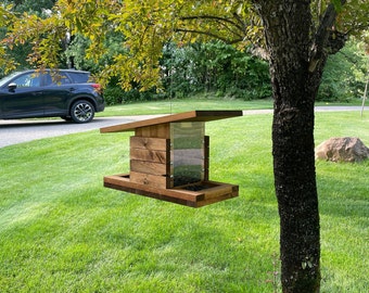 May include: A wooden bird feeder with a modern design, hanging from a tree branch. The feeder has a rectangular shape with a clear plastic viewing area and a sloped roof. The wood has a natural, warm brown tone. The feeder is suspended by a thin wire.