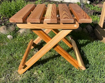 May include: A wooden folding stool with a natural wood finish. The stool is made of multiple slats of wood and has a simple, rustic design.