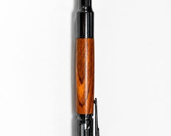 May include: A unique pen designed to resemble a bullet. The pen features a copper-colored tip, a black metal casing, a wooden barrel with a rich, brown grain, and a bolt-action clip. The pen is a distinctive writing instrument.