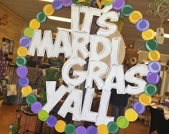 Może przedstawiać: Biały drewniany znak z napisem "It's Mardi Gras Y'all" w kółku otoczonym fioletowymi, zielonymi i złotymi koralikami. Znak jest zawieszony na sznurku i ma fioletową i złotą kokardkę na górze.