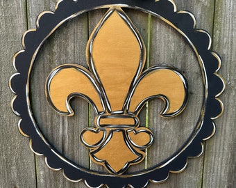 May include: A black and gold fleur-de-lis door hanger with a black scalloped frame. The fleur-de-lis is outlined in black and has a gold finish. A black and gold polka dot bow is attached to the top of the hanger.