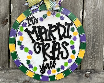 May include: Round wooden Mardi Gras sign with a white center and a colorful border of green, purple, and yellow. The sign reads "It's Mardi Gras y'all" in black lettering and is decorated with a festive bow.