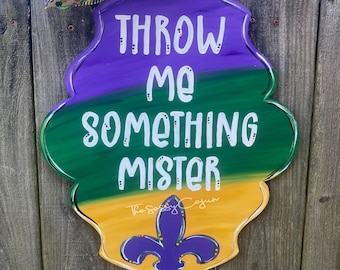 May include: A purple, green, and gold Mardi Gras door hanger with the text "Throw Me Something Mister" and a fleur-de-lis design. The door hanger is decorated with beads and a purple bow with white polka dots.