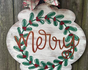 May include: A white wooden Christmas door hanger with a green holly wreath and the word "Merry" in red and gold lettering. A plaid and burlap bow with white lace is attached to the top.