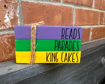 Może przedstawiać: Trzy drewniane klocki ułożone jeden na drugim. Górny klocek jest fioletowy i napisane jest na nim "BEADS". Środkowy klocek jest zielony i napisane jest na nim "PARADES". Dolny klocek jest żółty i napisane jest na nim "KING CAKES".