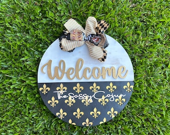 May include: A round wooden welcome sign with a black and gold fleur-de-lis pattern. The top half of the sign is white with the word "Welcome" in gold lettering. A gold and black bow is attached to the top of the sign. The text "The Sassy Cajun" is written in gold lettering on the bottom half of the sign.