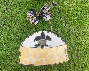 May include: A white and gold door hanger with a black fleur-de-lis design and the text "The Sassy Cajun".