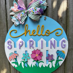 May include: A round, light blue wooden sign with the words "hello SPRING" in yellow and purple. The sign features a white picket fence with colorful flowers and a multi-colored bow at the top. The sign is hanging on a wooden fence.