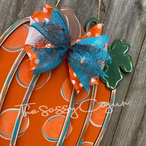 Pu&ograve; includere: Appendiporta a forma di zucca arancione con pois color teal e bianco e un fiocco color teal, bianco e arancione. La zucca ha una foglia verde sul lato. Il testo "The Sassy Cajun" &egrave; visibile al centro in basso dell'immagine.
