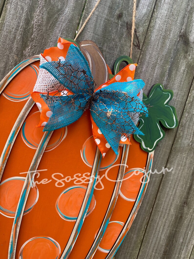 Pu&ograve; includere: Appendiporta a forma di zucca arancione con pois color teal e bianco e un fiocco color teal, bianco e arancione. La zucca ha una foglia verde sul lato. Il testo "The Sassy Cajun" &egrave; visibile al centro in basso dell'immagine.