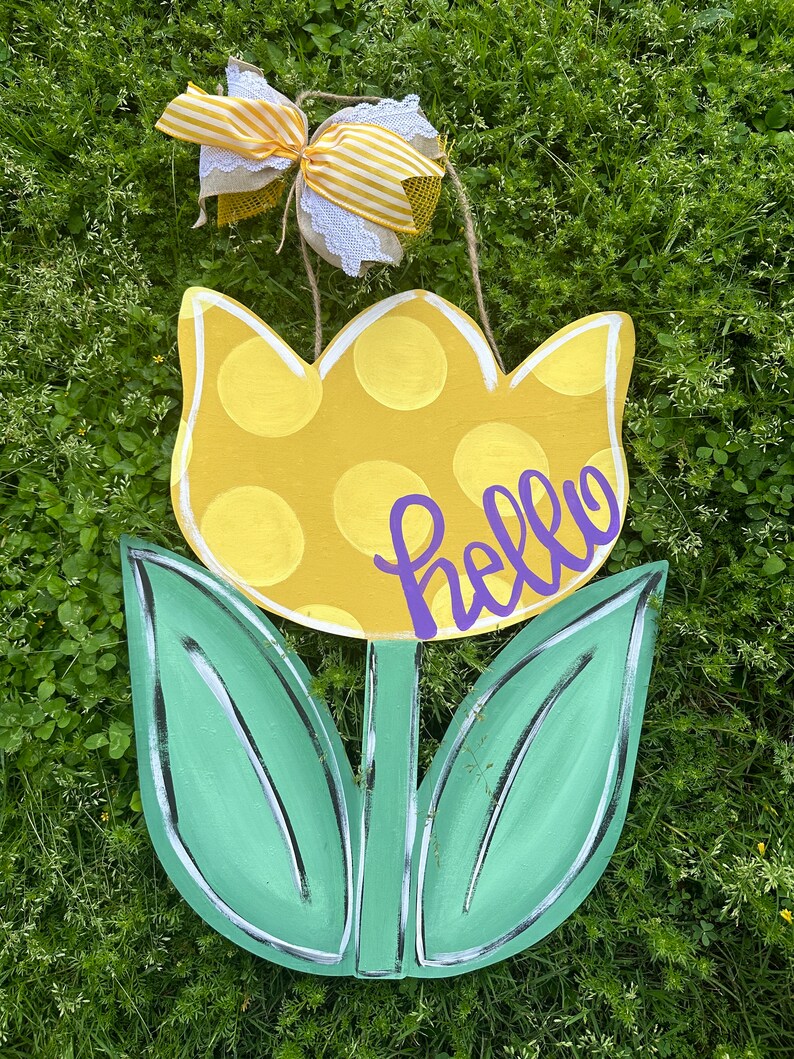K&ouml;nnte beinhalten: Ein h&ouml;lzernes tulpenf&ouml;rmiges Schild mit einer gelben, gepunkteten Blume und gr&uuml;nen Bl&auml;ttern. Das Wort "hello" ist in lila Schrift geschrieben. Oben ist eine dekorative Schleife mit gelb-wei&szlig; gestreiftem Band und Spitze befestigt.