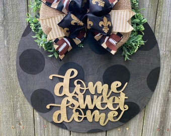 May include: A black and gold "home sweet home" sign with a black and gold bow with fleur-de-lis and football designs. The sign is decorated with black polka dots and is hanging on a wooden fence.