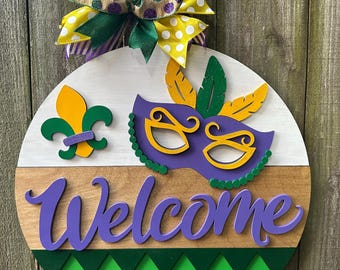 May include: A round wooden door hanger with a purple, yellow, and green Mardi Gras mask, a fleur-de-lis, and the word "Welcome" in purple lettering. The door hanger has a green and white diamond pattern at the bottom and is decorated with a purple, green, and yellow bow.