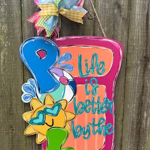 May include: A pink and orange door hanger with the text "Life is better by the" and a blue letter "P" with a yellow sun and a beach ball. The door hanger is decorated with a colorful bow.