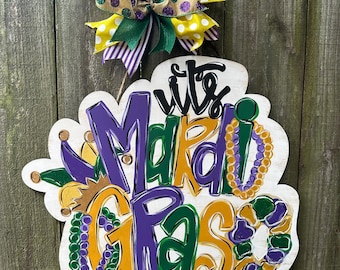 May include: A wooden Mardi Gras sign with the words "It's Mardi Gras Y'all" in purple, green, and gold. The sign is decorated with a bow in yellow, green, and purple. The sign is hanging on a wooden fence.