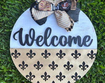 May include: A round wooden welcome sign with a black and white striped bow and a black and gold fleur-de-lis pattern. The sign says "Welcome" in black letters.