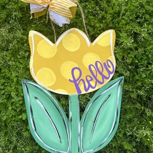 K&ouml;nnte beinhalten: Ein T&uuml;rh&auml;nger in Tulpenform aus Holz. Die Blume ist gelb mit wei&szlig;en Punkten, die Bl&auml;tter sind gr&uuml;n. Das Wort "hello" ist in lila Schrift geschrieben. Eine dekorative Schleife mit gelb-wei&szlig; gestreiftem Band ist befestigt.