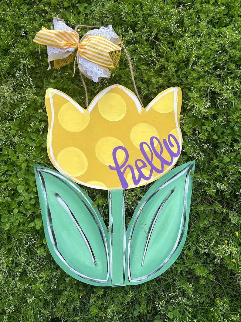 K&ouml;nnte beinhalten: Ein T&uuml;rh&auml;nger in Tulpenform aus Holz. Die Blume ist gelb mit wei&szlig;en Punkten, die Bl&auml;tter sind gr&uuml;n. Das Wort "hello" ist in lila Schrift geschrieben. Eine dekorative Schleife mit gelb-wei&szlig; gestreiftem Band ist befestigt.
