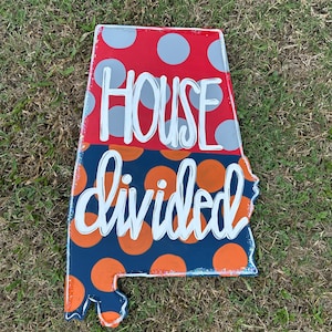 May include: A wooden sign shaped like the state of Alabama with a red and white polka dot pattern on the top half and a blue and orange polka dot pattern on the bottom half. The sign says "HOUSE divided" in white lettering.