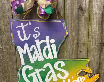 Może przedstawiać: Drewniany znak w kształcie stanu Luizjana z efektem ombre w kolorze fioletowym, zielonym i żółtym. Znak głosi "It's Mardi Gras y'all" białą farbą. Fioletowa, zielona i złota kokarda z białymi kropkami wisi nad znakiem.