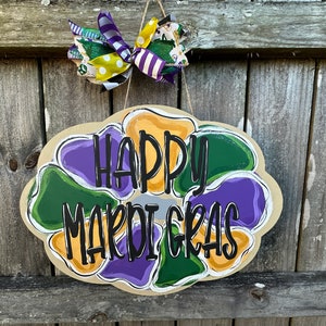 May include: A round, brown wooden door hanger with a purple, green, and yellow flower design. The words "Happy Mardi Gras" are written in black lettering on the door hanger.