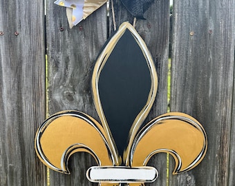 May include: A black and gold fleur-de-lis wooden door hanger with a white stripe across the center. The hanger is decorated with a black and gold bow.