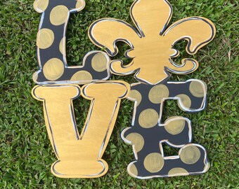 May include: A wooden "LOVE" sign with a fleur-de-lis in the center. The letters are painted black with gold polka dots and outlined in white. The fleur-de-lis is gold with white accents.