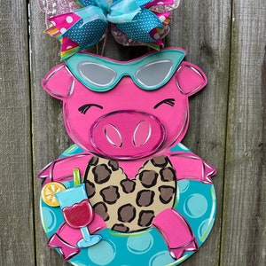 May include: A pink pig wearing sunglasses and a leopard print swimsuit is floating in a blue and white inflatable ring. The pig is holding a pink drink with a lime wedge.