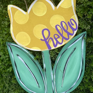 K&ouml;nnte beinhalten: Ein h&ouml;lzernes tulpenf&ouml;rmiges Schild mit einem gelben, gepunkteten Bl&uuml;tenkopf und gr&uuml;nen Bl&auml;ttern. Das Wort "hello" ist in lila Schrift &uuml;ber der Blume geschrieben. Die R&auml;nder sind wei&szlig; umrandet.